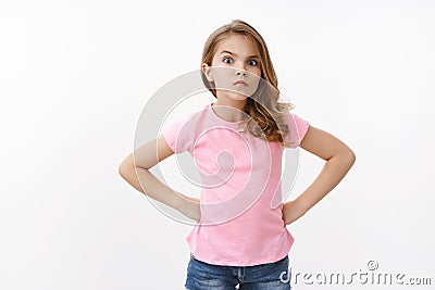 Serious-looking strict and angry little arrogant girl, blond child, stand in bossy offended pose, hold hands waist Stock Photo