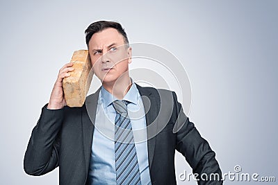 A serious man in a jacket and tie talks with a yellow brick pressed to his ear. On a light blue background. Stock Photo