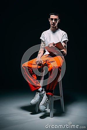 Serious man in orange uniform with cuffs on legs reading bible and sitting on stool Stock Photo