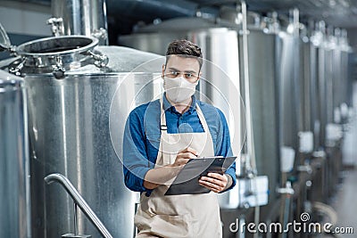 Serious man standing in brewery and write down data of equipment during covid-19 quarantine Stock Photo