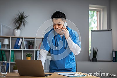 Serious millennial chinese male doctor calling by phone and looking at laptop in clinic office interior Stock Photo
