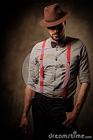 Serious old-fashioned man with hat wearing suspenders and bow tie, posing on dark background. Stock Photo