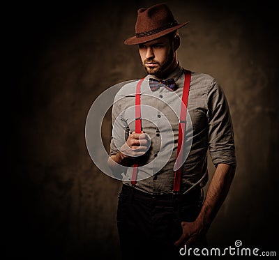 Serious old-fashioned man with hat wearing suspenders and bow tie, posing on dark background. Stock Photo