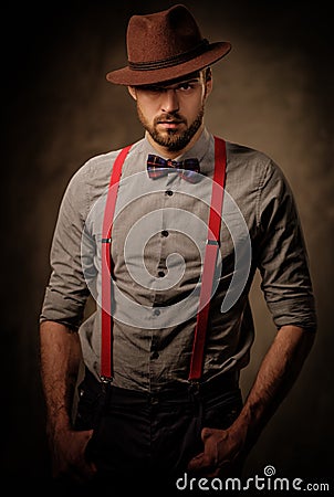 Serious old-fashioned man with hat wearing suspenders and bow tie, posing on dark background. Stock Photo