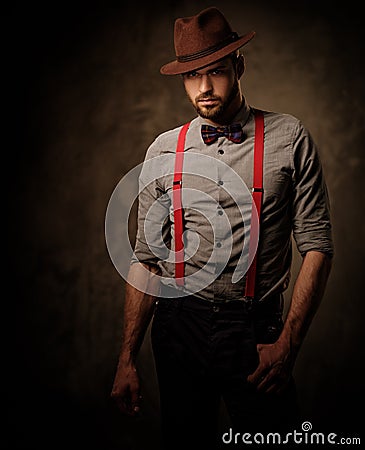 Serious old-fashioned man with hat wearing suspenders and bow tie, posing on dark background. Stock Photo