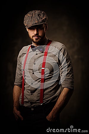 Serious old-fashioned man in tweed hat wearing suspenders and bow tie, posing on dark background. Stock Photo