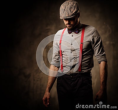 Serious old-fashioned man in tweed hat wearing suspenders and bow tie, posing on dark background. Stock Photo