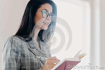 Serious thoughtful brunette European woman writes down text information in notepad, concentrated somewhere into distance, makes Stock Photo