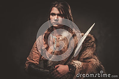 Serious viking woman with sword in a traditional warrior clothes, posing on a dark background. Stock Photo