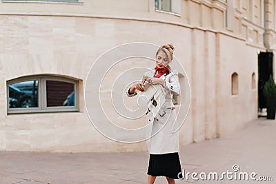 Serious woman in long beige coat walking by building and reading fresh news. Unhappy blonde lady in black dress hurries Stock Photo