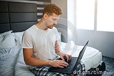 Serious young man in bed this morning. He work at home. Guy type on laptop keyboard and look at screen. Daylight. Stock Photo