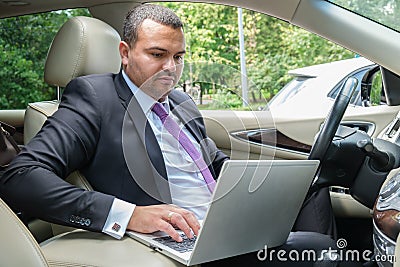 Serious young man in a business suit sits in the driver`s seat in the car with laptop. Busy businessman with short hair Stock Photo
