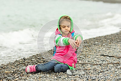 Seven-year girl sitting on a pebble beach in warm clothing and pours out through her fingers small stones Stock Photo