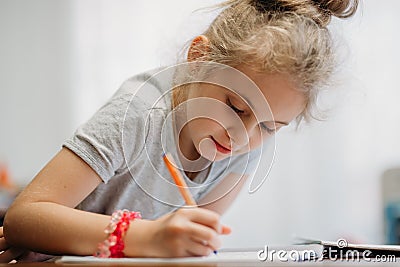 A seven-year-old girl sits at home at a table and writes in a notebook, completing a learning task or repeating lessons. Stock Photo