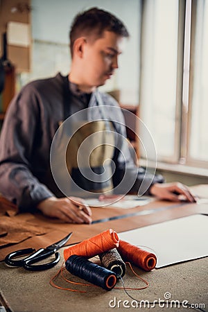 Several spools of colored thread on the desktop of a tailor, who is in the background in out-of-focus sewing a leather Stock Photo