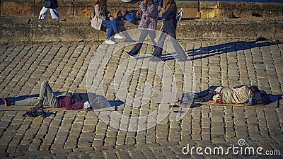 Seville, Spain April 7 2023: A world of contrasts. Black homeless people lying on the pavement while other people stroll or sit Editorial Stock Photo