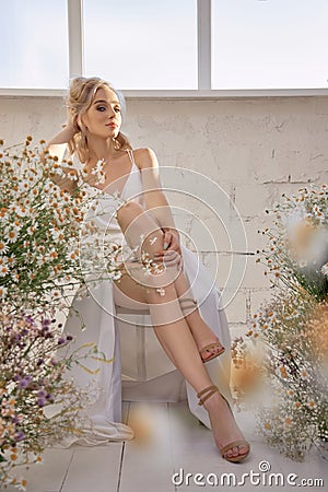 Sexy blonde woman in a beautiful white dress is sitting near the window in front of a bouquet of wild flowers. Romantic girl with Stock Photo