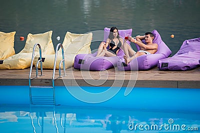 couple relaxing with drinks on cushioned loungers by swimming pool and river on the background Stock Photo