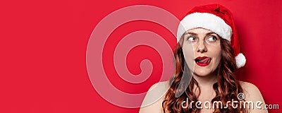 Sexy girl with red santa hat with pompom licks lips on red wall background Stock Photo