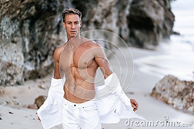 Sexy muscular guy with bare-chested in white pants takes off white shirt on beach, rocks on background Stock Photo