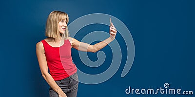 Sexy slim blonde womanin red t-shirt on a blue studio background making selfie with smartphone. Copyspace Stock Photo