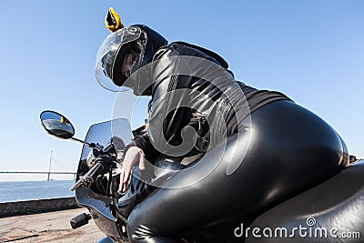Sexy woman motorcyclist in black leather apparel and helmet sitting on bike, rear view, low angle view Stock Photo