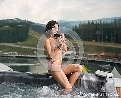 woman with perfect figure in bikini sitting on the Jacuzzi lowered feet in the water and drinking cocktail Stock Photo