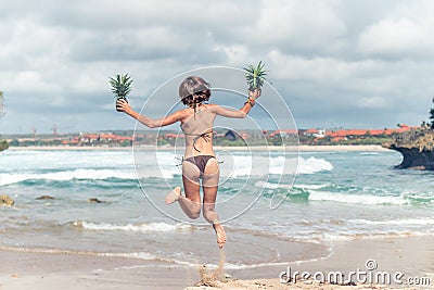 young lady in bikini jumping on the beach with fresh raw healthy pineapple fruit. Happy vacation concept. Bali. Stock Photo