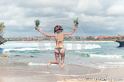 young lady in bikini jumping on the beach with fresh raw healthy pineapple fruit. Happy vacation concept. Bali. Stock Photo
