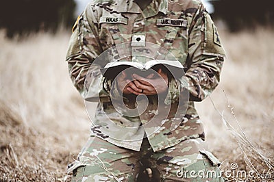 Shallow focus shot of a young soldier kneeling while holding a bible in a field Stock Photo