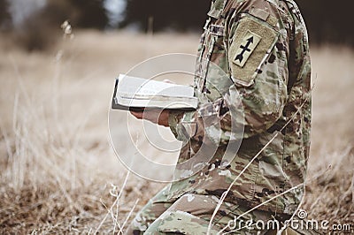 Shallow focus shot of a young soldier kneeling while holding an open bible in a field Stock Photo