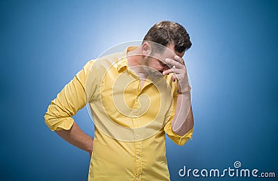Shame. Half length young man covering face with hands over blue background, dresses in yellow shirt. Oops. Ashamed human Stock Photo