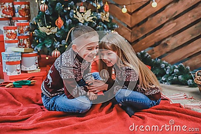 Sweet shot of a boy telling a secret in the ear to his sister sitting on red blanket at home, celbrating Christmas. Stock Photo