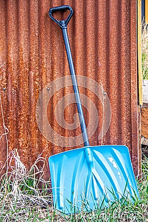Shinny blue metal show shovel leaning against a grainy rusty corregated shed with green and dry grasses growing at the bottom Stock Photo