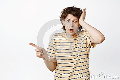 Shocked and anxious young man, pointing finger left, hold hand with troubled face, scared of something bad, standing Stock Photo