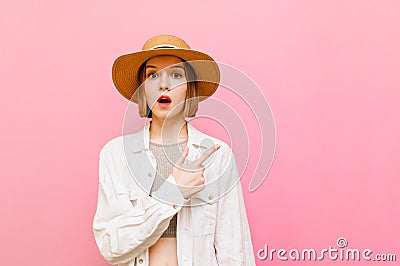 Shocked attractive girl stands against a pink background in a hat and white summer clothes,looks in camera and points finger to Stock Photo