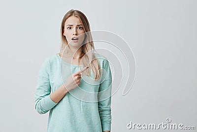 Shocked blonde girl in blue sweater with opened mouth frowning face looking in bewilderment at camera, pointing finger Stock Photo