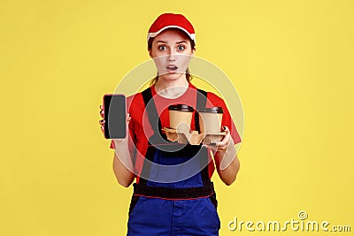 Shocked delivery woman holding take away coffee, showing mobile phone with blank screen. Stock Photo