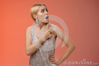 Shocked displeased bothered arrogant blond glamour woman in silver glittering dress turning upper right corner pointing Stock Photo