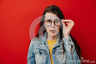 Shocked emotional young woman keeps hand on rim of spectacles, dressed in denim jacket, gazes with stupefaction Stock Photo