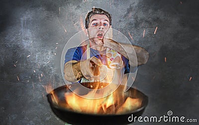 Shocked messy man with apron holding pan in fire burning the food in kitchen disaster and unskilled and unexperienced terrible ho Stock Photo