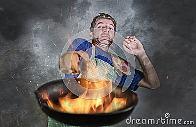 Shocked messy man with apron holding pan in fire burning the food in kitchen disaster and unskilled and unexperienced terrible ho Stock Photo