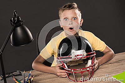 shocked schoolboy sitting with american football helmet at table with eyeglasses lamp colour pencils and books on grey Stock Photo