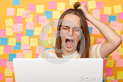 Shocked worried woman with brown hair working on laptop against yellow wall with memo cards, having problems with her online work Stock Photo