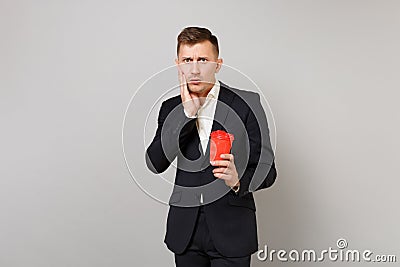 Shocked young business man in classic black suit putting hand on cheek holding paper cup with coffee or tea isolated on Stock Photo