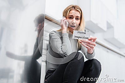 Shocked young woman holding card with lettering home inspection and talking on smartphone near broken refrigerator Stock Photo