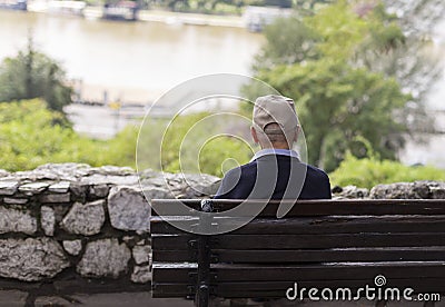 A lonely old man sitting on a bench in a park, looking at river Stock Photo