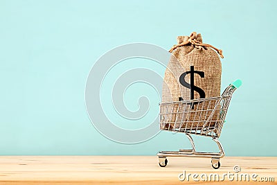 shopping cart with bag full of money with dollar sign over blue wooden background. Stock Photo