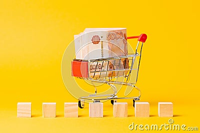 A shopping cart filled with a five-thousand-ruble bill. There are wooden cubes for writing the word. Yellow background. Close up. Stock Photo