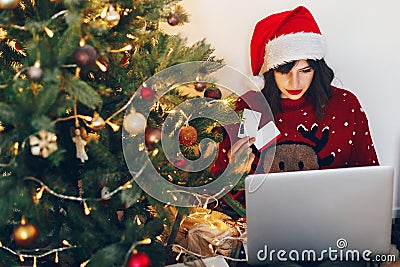 shopping online and christmas sale concept. stylish woman in reindeer hat holding credit card and laptop, under christmas tree li Stock Photo